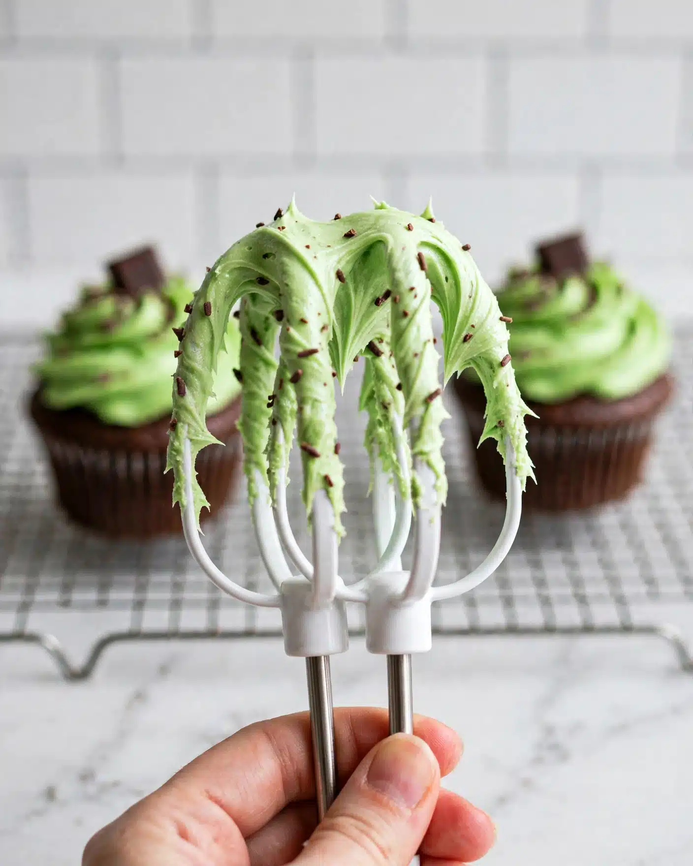 Mint Chocolate Cupcakes Recipe 4 A close-up image shows a white mixer beater covered in light green frosting with a creamy, smooth, and swirled texture. A woman's hand holds the beater from the bottom, with the background featuring two chocolate cupcakes in white paper liners on a metal rack against a white marbled surface and white subway tile. The image is bright and clean, focusing on the soft, thick frosting on the beater photo taken with an iphone --ar 4:5 --v 7