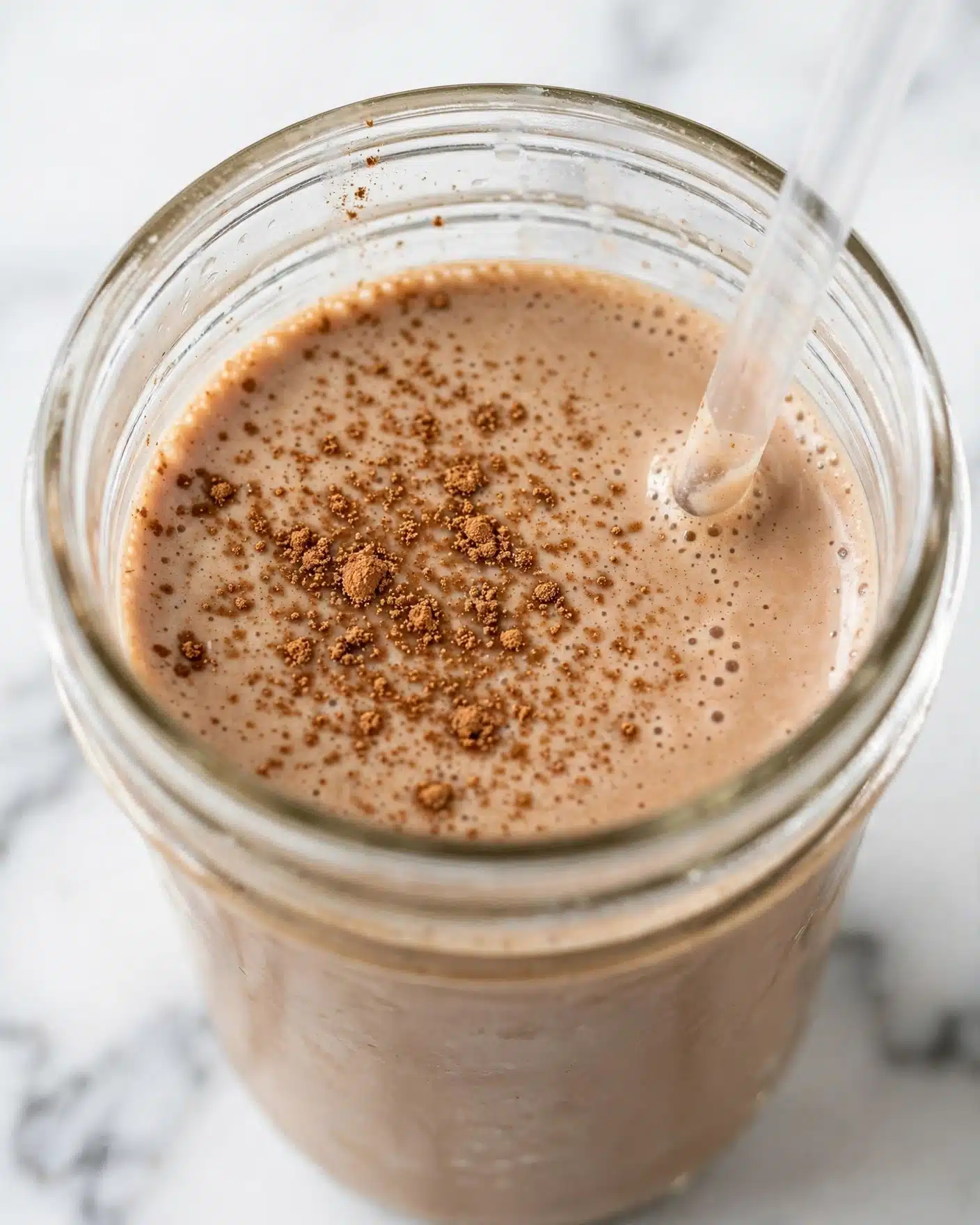 Pumpkin Protein Shake Recipe 5 The image shows a close-up of a jar filled with a light brown creamy drink topped with scattered darker brown powder, likely cinnamon or nutmeg. The jar is clear glass, revealing the smooth texture of the drink inside, and there is a clear straw inserted on the right side. The background and surface are a white marbled texture, adding a clean and bright feel to the scene. The focus is on the top of the jar and the drink's surface, capturing the tiny bubbles and powder details clearly. photo taken with an iphone --ar 4:5 --v 7