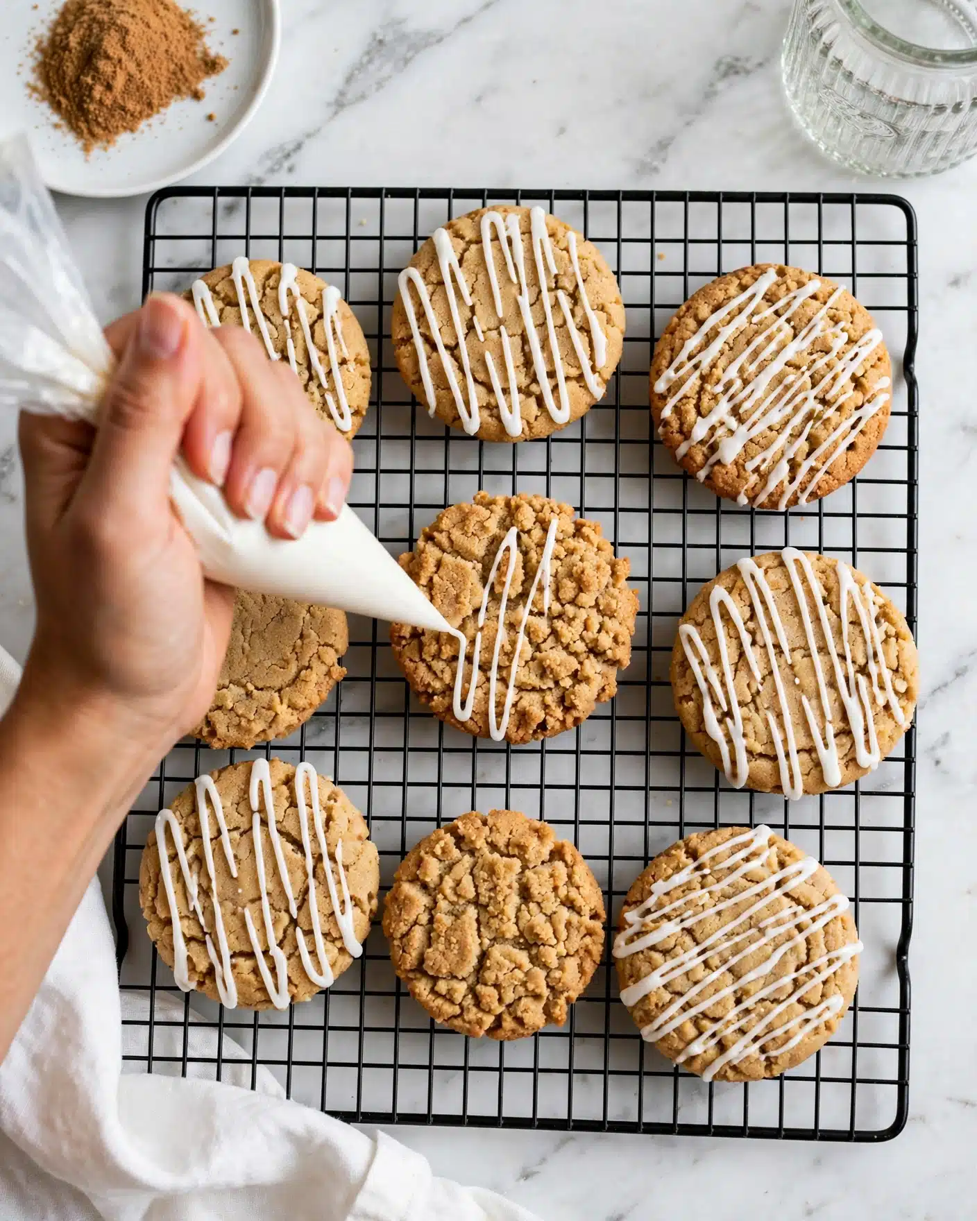 Coffee Cake Cookies with Streusel Topping and Vanilla Icing Recipe 5 There are eight round cookies with a golden brown color and crumbly texture on a black wire rack, all placed on a white marbled surface. Six of the cookies have thin white icing lines drizzled over the top in a neat pattern, while two cookies are plain, showing their crumbly, rough tops. A woman's hand is holding a white piping bag, delicately adding white icing drizzle onto one of the plain cookies in the center. The top left corner shows a white plate with some brown powder, and part of a textured glass jar is visible near the top right. A white cloth is partially visible at the bottom left. Photo taken with an iphone --ar 4:5 --v 7