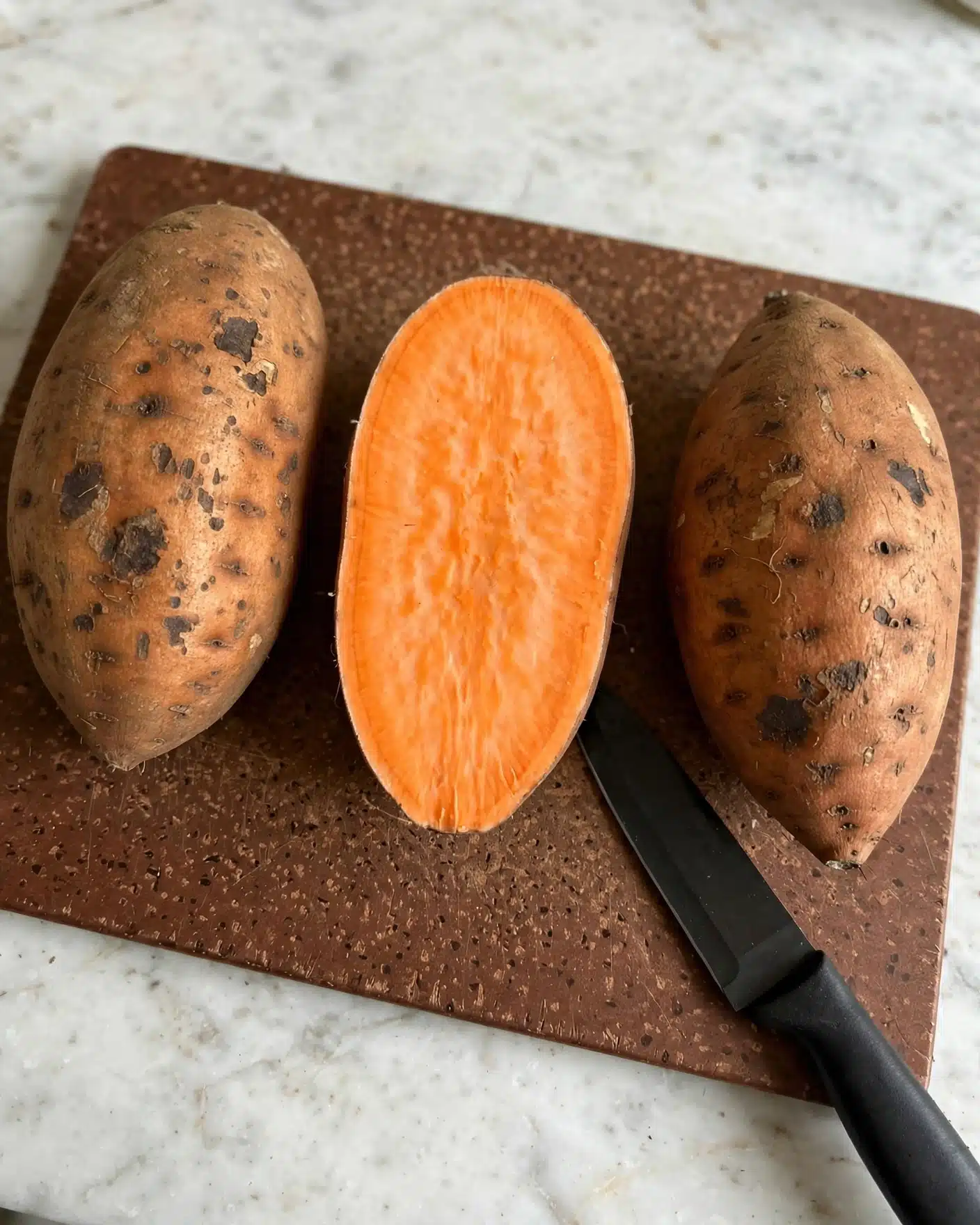 Lightened Up Sweet Potato Casserole Recipe 4 The image shows two whole sweet potatoes and one sweet potato cut in half, all placed in a row on a brown cutting board with a textured surface. The two whole sweet potatoes have a rough, brownish-orange skin with dark spots, while the cut sweet potato reveals a smooth, bright orange inside with subtle light streaks. A knife with a black blade lies near the cut sweet potato on the cutting board. The cutting board is set on a white marbled surface. photo taken with an iphone --ar 4:5 --v 7