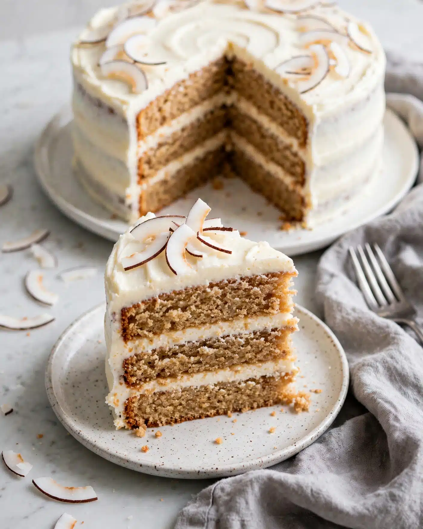 A slice of cake with three layers sits on a small speckled plate; the bottom and middle layers are light brown and crumbly while the two creamy white frosting layers separate and top the cake. Thin, curved white coconut flakes with dark edges decorate the top layer of frosting. Behind the slice, a larger white plate holds the rest of the cake, showing the same three-layer pattern with the white frosting covering the sides. The setting is on a white marbled surface with a grey cloth and a silver fork slightly visible nearby. Photo taken with an iphone --ar 4:5 --v 7