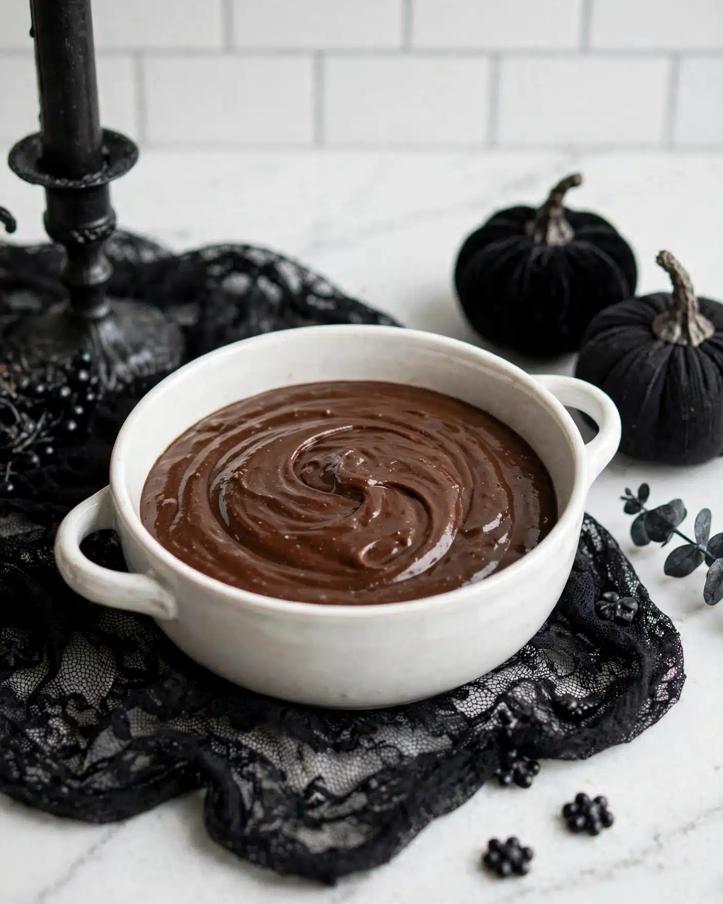 Black Velvet Cupcakes with Black Cocoa Buttercream Recipe 4 A round white ceramic bowl with two small handles holds a smooth, dark brown chocolate batter with light swirls visible on the surface. The batter fills almost the whole bowl, showing a glossy texture. The bowl sits on a black lace cloth on a white marbled surface. Around the bowl, there are black decorative items including a candle holder and fabric pumpkins, giving a cozy and slightly spooky feeling. Photo taken with an iphone --ar 4:5 --v 7