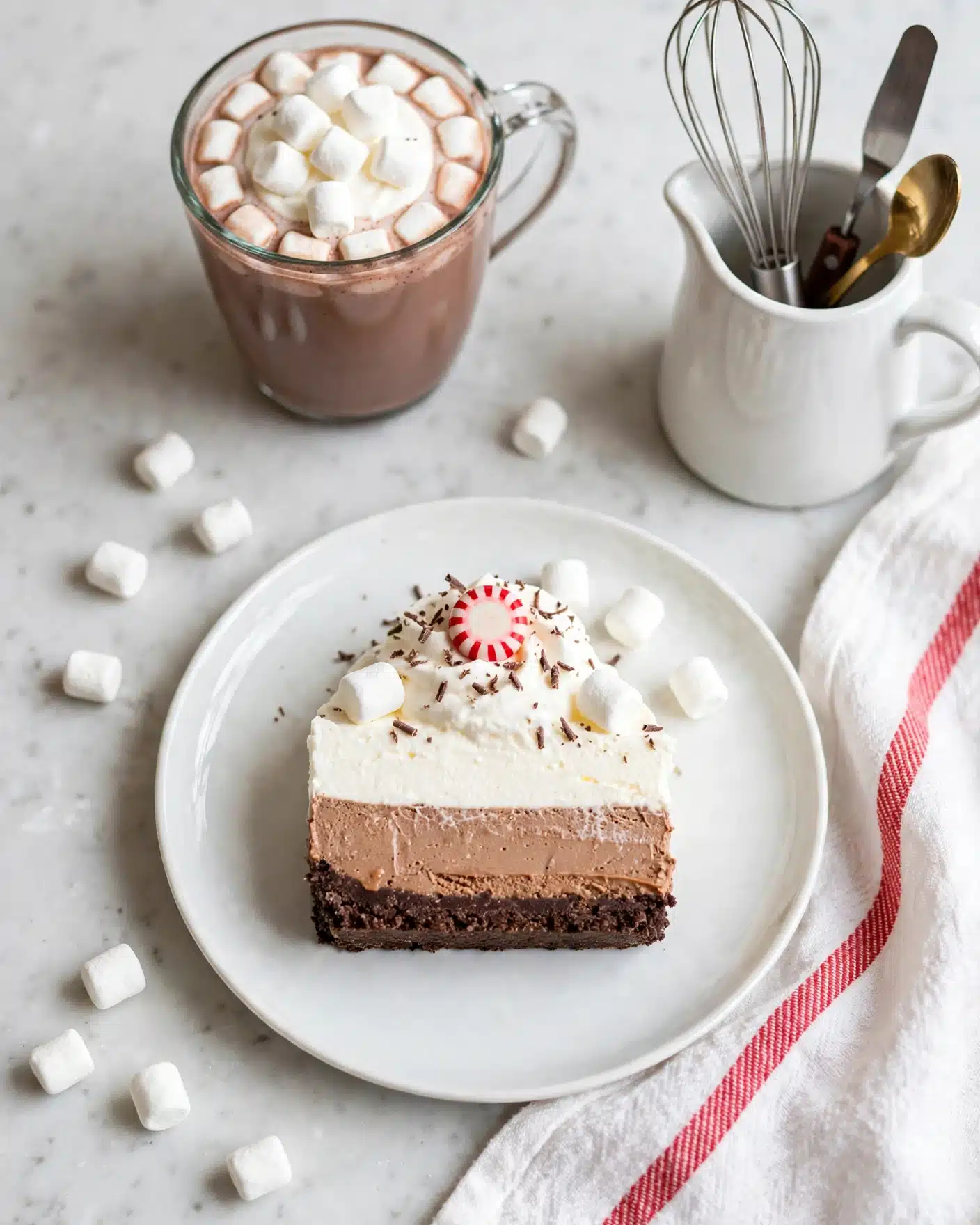No Bake Peppermint Hot Chocolate Cheesecake Recipe 5 A slice of layered dessert sits on a white plate over a white marbled surface covered partially by a white cloth with a red stripe. The dessert has three layers: a dark brown bottom crust, a thick light brown middle layer, and a thick white top layer. On the top layer, there is white whipped cream with a small red and white striped peppermint candy in the center, small white marshmallows, and chocolate shavings scattered around. More marshmallows surround the plate. In the background, there is a glass cup filled with hot chocolate topped with mini white marshmallows and a white pitcher holding kitchen utensils. Photo taken with an iphone --ar 4:5 --v 7
