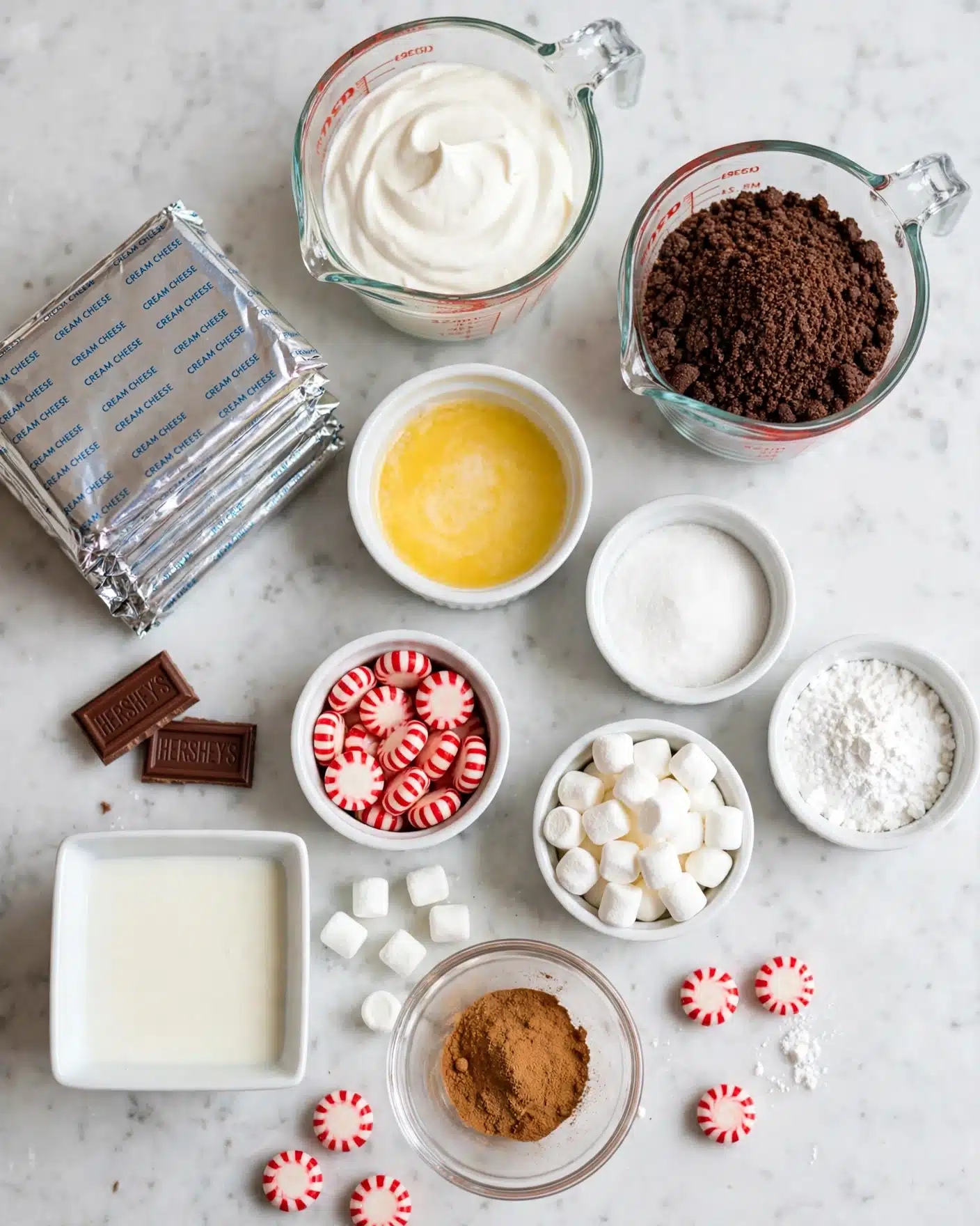 No Bake Peppermint Hot Chocolate Cheesecake Recipe 4 The image shows multiple small white bowls and a few glass measuring cups arranged on a white marbled surface. One clear glass measuring cup at the top center holds white cream. To its right, a glass measuring cup is filled with dark brown cookie crumbs. Below the cream is a white ramekin with bright yellow melted butter. Surrounding these are several white ramekins filled with fine white sugar, coarse white powder, small white marshmallows, and red-white peppermint candies. An additional clear bowl holds light brown cinnamon powder. At the bottom left, there are two small pieces of Hershey's chocolate bars and a small white square bowl with a light liquid. Several foil-wrapped rectangular blocks, likely cream cheese, are stacked on the left side. The shot has bright, even lighting, and the clean simple arrangement emphasizes the varied colors and textures of the ingredients. Photo taken with an iphone --ar 4:5 --v 7