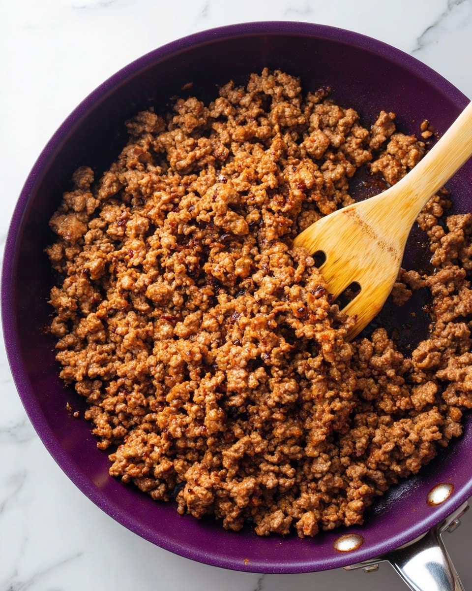 The image shows a close-up of browned ground meat in a deep purple pan, with small pieces cooked evenly to a light and dark brown color with some crispy bits. A wooden spatula is stirring the meat from the top right corner, with the texture looking crumbly and moist. The pan is placed on a white marbled surface, adding brightness to the setting photo taken with an iphone --ar 4:5 --v 7