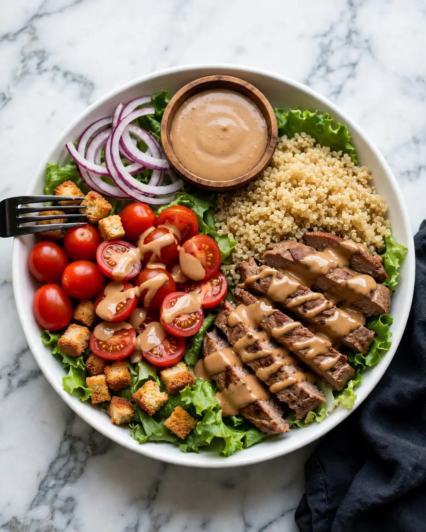 A white bowl with a base layer of green leafy lettuce at the bottom edges is topped with sliced red onions on one side and a serving of cooked quinoa on the opposite side. Next to the quinoa are sliced grilled meat pieces with a brown color and a drizzle of creamy light brown sauce over them. On the left side of the bowl, bright red cherry tomatoes, some halved and covered lightly with sauce, are placed above crispy golden-brown croutons scattered around. In the center near the top edge, there is a small wooden bowl filled with thick creamy light brown dressing. A black fork rests on the rim of the bowl next to the cherry tomatoes. The bowl sits on a white marbled surface with a piece of dark cloth nearby. photo taken with an iphone --ar 4:5 --v 7