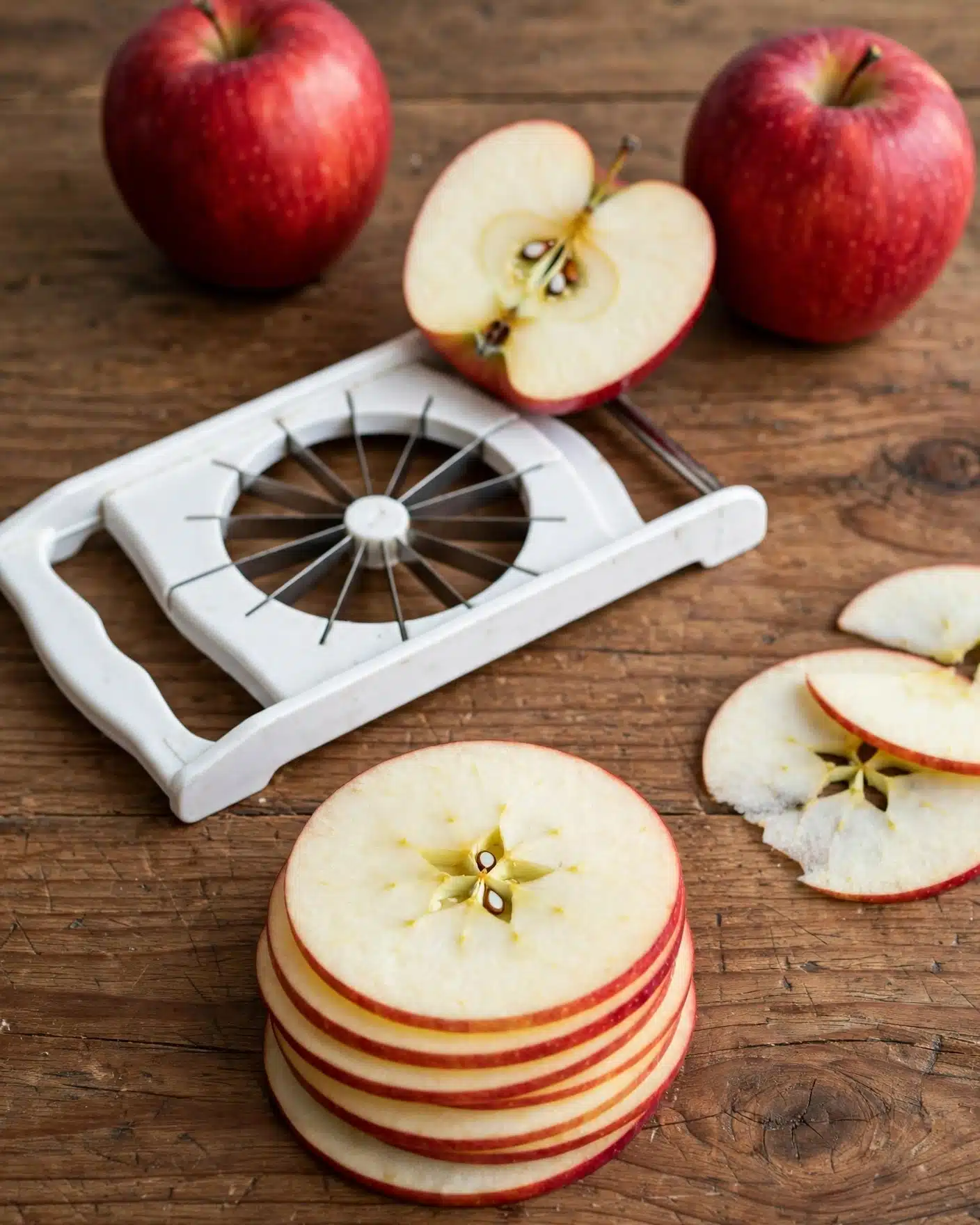 Easy Apple Chips Recipe 4 The image shows a white apple slicer on a brown wooden surface with whole and sliced red apples around it. There are two whole apples in the background, a halved apple with seeds visible towards the center, and neatly stacked thin apple slices with a slight red border in the foreground. The slices reveal the star-shaped core pattern clearly, and the wooden surface’s grain is visible under the fruit and slicer. Photo taken with an iphone --ar 4:5 --v 7