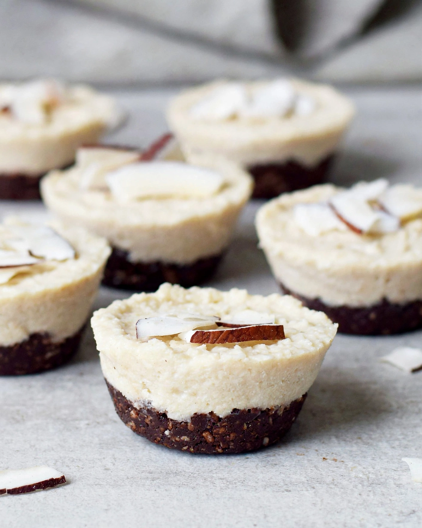 Vegan Coconut Cups Recipe 5 The image shows six small dessert cups arranged on a white marbled surface. Each dessert has two layers; the bottom layer is dark brown, crumbly, and textured, resembling a chocolate or nut base. The top layer is thick, creamy, and pale beige, with a soft, slightly grainy texture. On top of each dessert, there are thin, flat slices of coconut garnish in white with brown edges. The focus is on the front dessert cup, with the others softly blurred in the background. Photo taken with an iphone --ar 4:5 --v 7